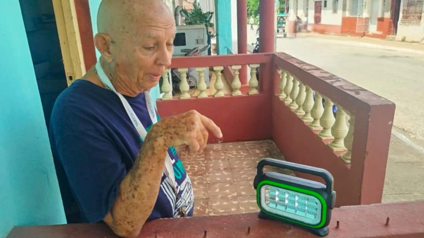 Older man in a blue shirt sits on a porch, holding a water bottle beside a green digital clock.