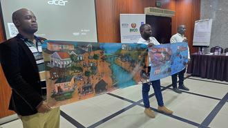 Three men hold large banners depicting vibrant scenes of a village at a conference room.