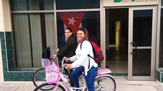 Two women pose on pink bicycles in front of a building with a Cuban flag.