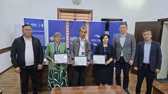 Seven people pose with certificates on a stage in a conference room, blue banners behind them.