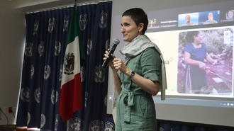 Person in green scrubs presenting on stage with microphone; projector screen and flags in background.