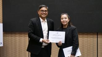 Two people on stage; man in a suit presents a certificate to a woman.