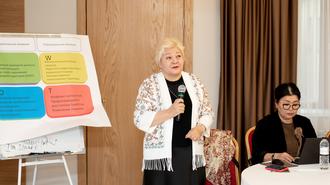 Photograph of a woman presenter with a white scarf speaking at a workshop; colorful chart on easel.