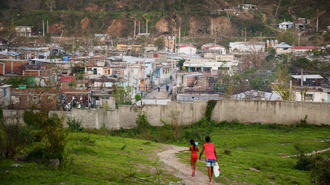 Two people walk a dog along a grassy path toward a hillside town of many small houses.