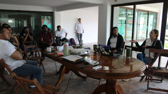 Photo: group of people seated around a round wooden table in a bright room.