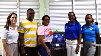 Five adults pose together in front of storefront shutters, with a blue box on a table.