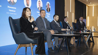 Panel of five speakers seated on stage behind a long table; blue backdrop with portraits.