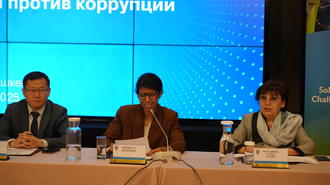 Three panelists seated at a conference table with microphones and water, blue backdrop.