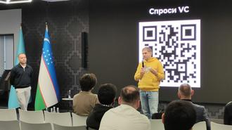 Audience watches a speaker in a yellow shirt on stage next to a large QR code and flags.