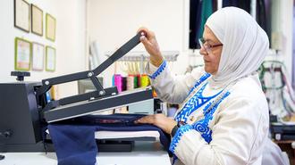 Person in a white hijab uses a large sewing machine to sew blue fabric in a bright studio.