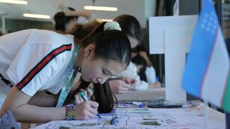 Woman leaning over a table, drawing on large papers with markers in a classroom; a flag nearby.