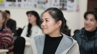 People seated in a classroom; woman in light gray cardigan and black top, posters on wall.