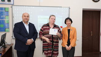 Three people in a classroom pose with a certificate; whiteboard, clock, and door behind.