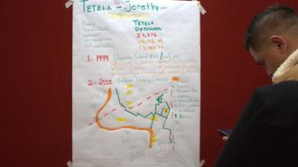 Man in a dark suit studies a large white poster with a colorful hand-drawn map on a red wall.