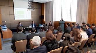 Panelists at a long table presenting to a seated audience in a conference room.