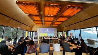 People sit around a long table in a bright room with large windows and a presentation on screen.