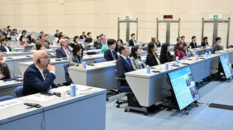 Conference hall filled with attendees seated at blue desks with screens.
