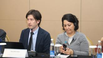 Two attendees sit at a conference table with microphones and water bottles.