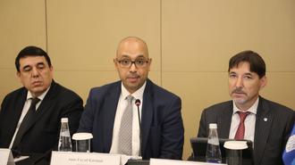 Three men in business suits sit at a conference table with nameplates and water glasses.