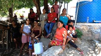 Group of people outdoors beside a stone wall with a blue water tank in the background.
