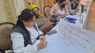 Photo of a woman writing on a large whiteboard at a workshop; others sit at a table in an ornate room.