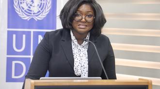 Photograph of a woman speaking at a UN podium, with a UN banner in the background.
