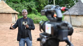 Man in a black jacket speaks to camera as filming gear looms in foreground, village huts behind.