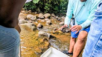 People by a rocky creek with a white cooler; one bends toward the water, another sits nearby.