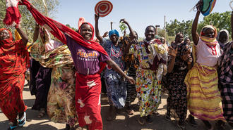 Group of women in bright traditional dresses dancing outdoors, raising arms with round fans.