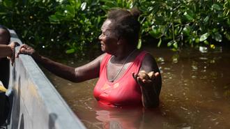 Woman in a red sleeveless top stands waist-deep in muddy water, gesturing beside a boat.