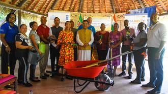 Diverse group stands around a red wheelbarrow beneath a thatched roof.