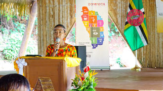Woman in a bright patterned shirt at a podium on stage, with banner and flowers.