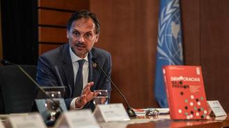Photograph of a suited man at a conference table with microphones, UN flag in the background.