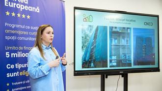 Female presenter in a light blue blouse beside a large monitor showing slides; blue banner left.