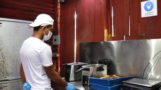 Photograph: male cook in white cap and mask by stainless steel kitchen counter with blue tray.