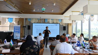 Workshop attendees in a conference room sit at tables as a presenter speaks at a projection screen.