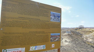 Outdoor wooden information sign with text blocks and a small photo, set in a dry desert landscape.