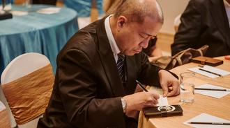 Bald man in a suit signs a document at a conference table.