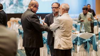 Photo shows two men in suits shaking hands at a formal event, teal banquet tables in background.