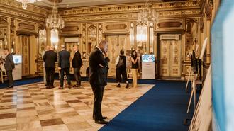 Ornate ballroom with gold decor and chandeliers; suited attendees stand on a blue carpet.