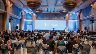 Blue-lit stage in a large conference hall with round tables and many attendees.