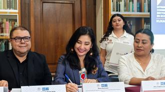 Four seated people at a signing event; a standing woman holds a document behind bookshelves.