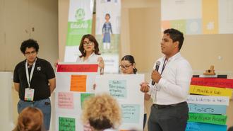 Speaker in a white shirt with a microphone speaks beside colorful project posters at a conference.