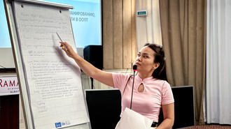 Photograph of a woman in a pink top pointing to a flip chart during a conference.