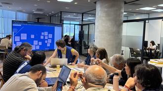 Photograph of a busy conference room with people around a long table and a blue screen in the background.