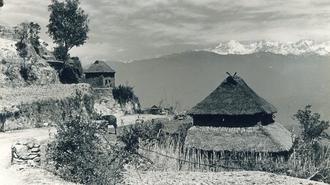 Black-and-white photo of hillside village with thatched round huts, stone walls, and distant mountains.