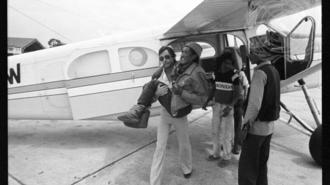 Black-and-white photo of a small propeller plane on the tarmac with people nearby.
