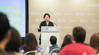 Speaker presenting on a conference podium, with a slide and an audience in the foreground.