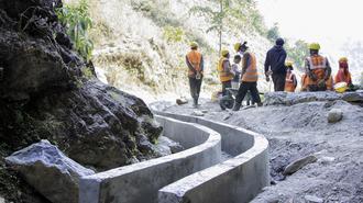 Construction workers in orange vests and hard hats inspect curved concrete barrier along a rocky road.