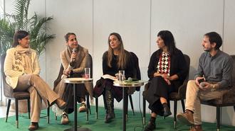 Five people sit on a panel on a green carpet, small round tables with cups.
